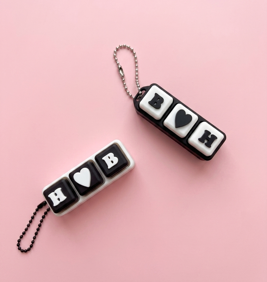 Two black and white keychains with heart and letter symbols on a pink background