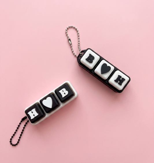 Two black and white keychains with heart and letter symbols on a pink background