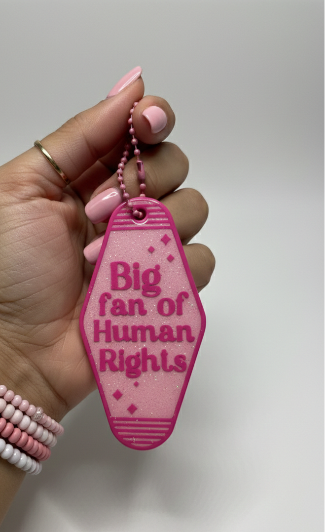 Hand holding a pink keychain with 'Big fan of Human Rights' text on a white background