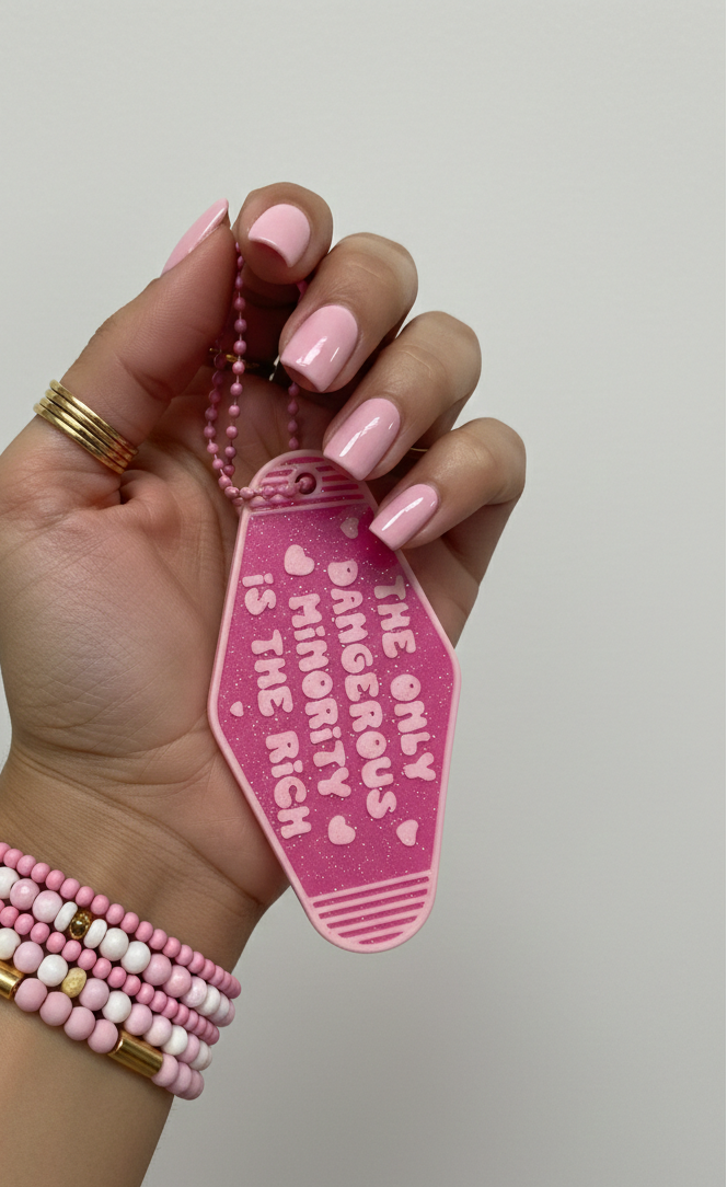 Hand wearing pink and gold bracelets holding a pink tag with text against a neutral background